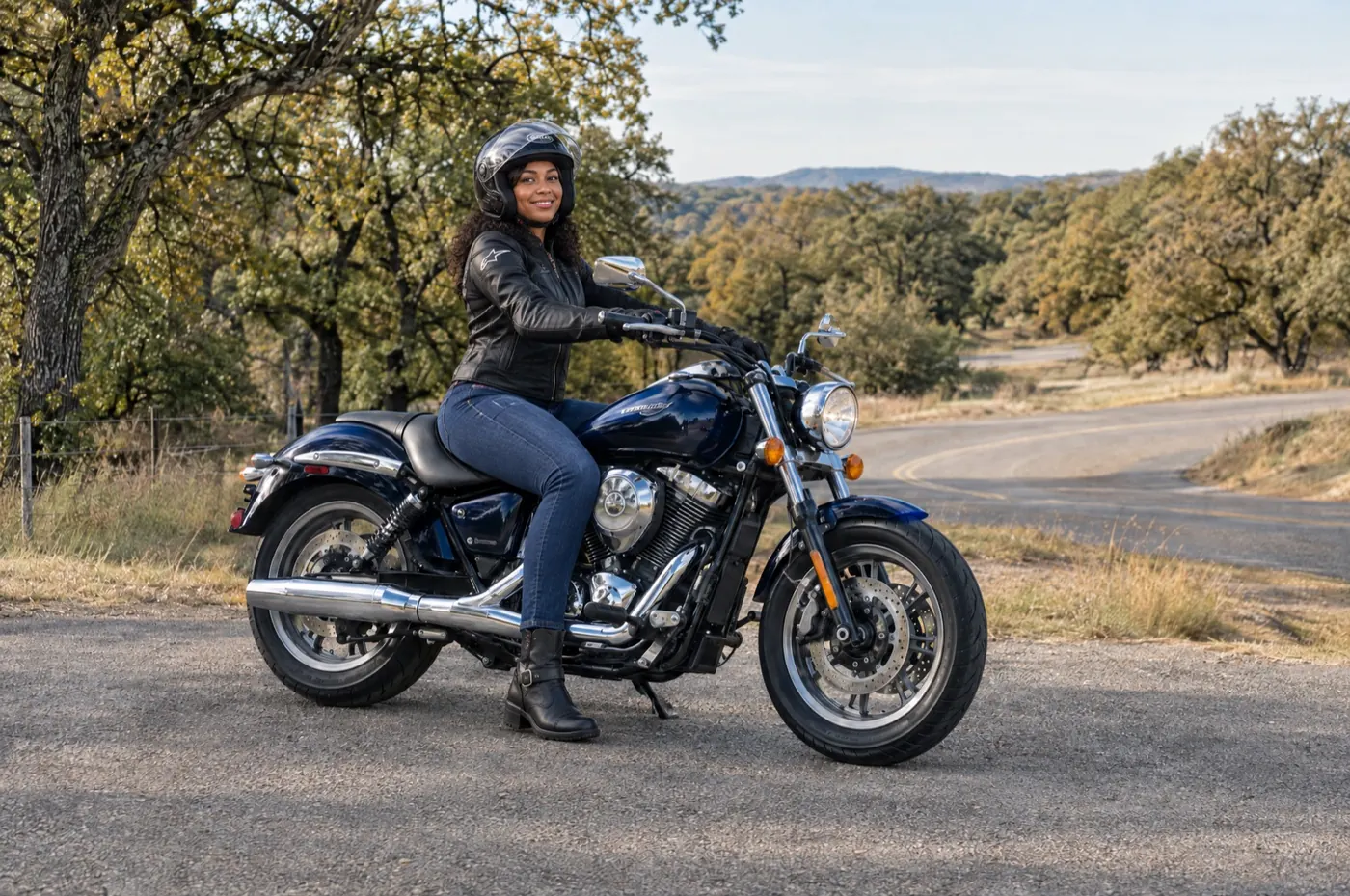 A woman wearing riding gear seated on a navy road cruiser beside a Texas country road